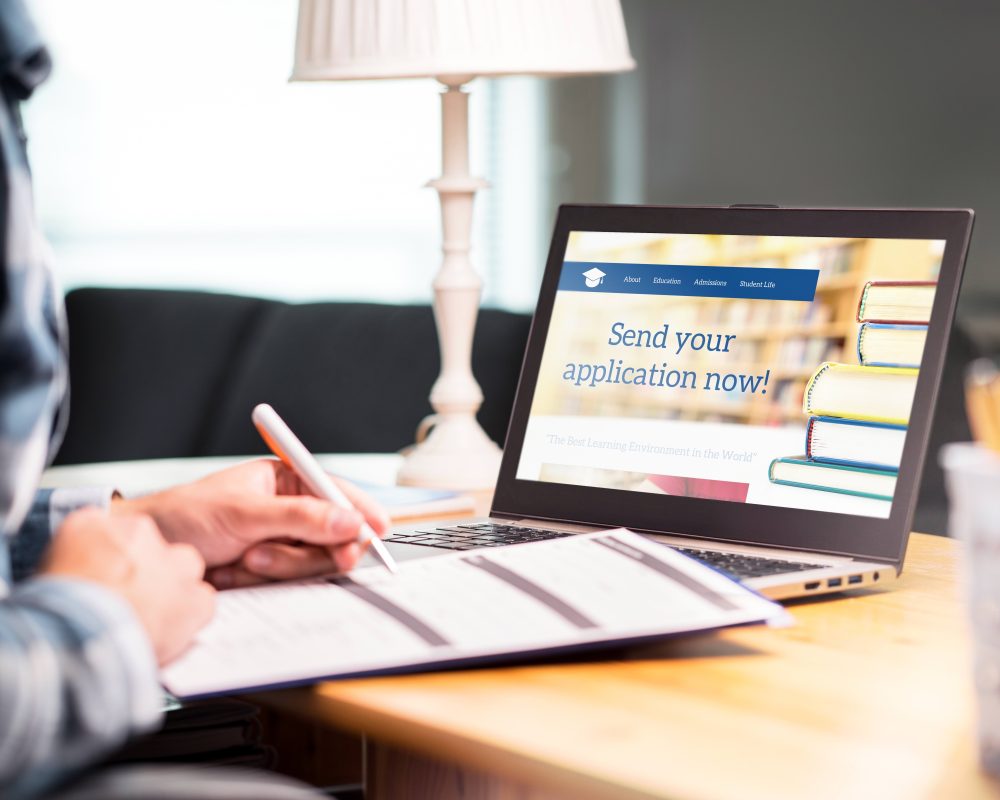 Young man writing college or university application form with pen and paper. Student applying or filling document. Scholarship, grant or admission. School website in laptop.