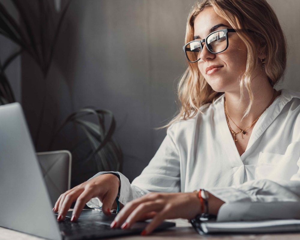 Happy young blonde business woman entrepreneur using computer looking at screen working in internet sit at office desk, smiling millennial female professional employee typing email on laptop at workplace